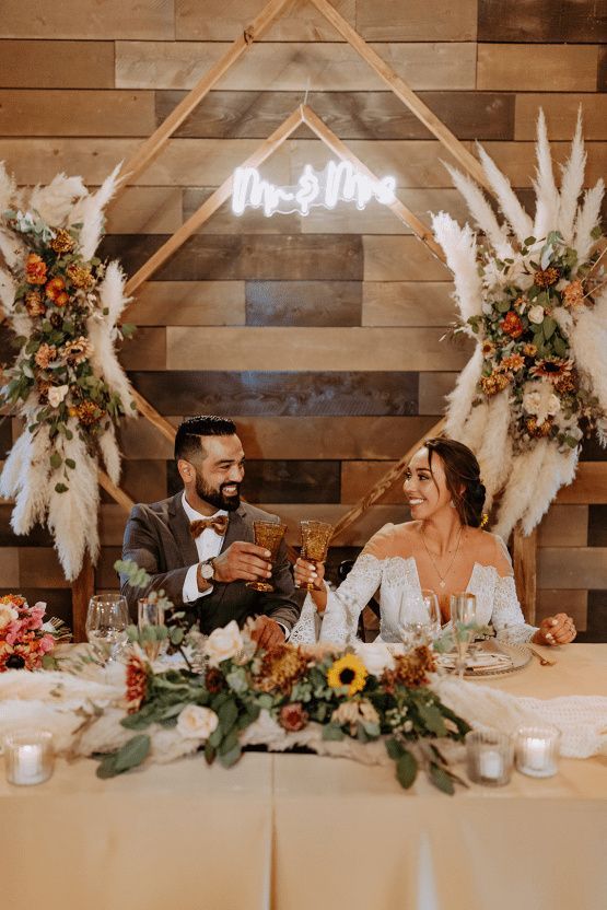 a boho wedding sweetheart table with greenery, neutral blooms and unsflowers, a neon sign and pampas grass with blooms in the back