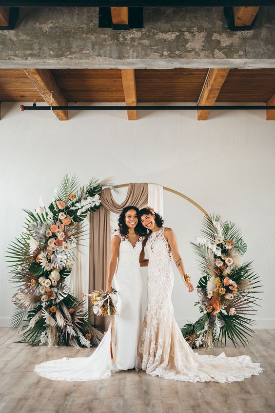 a creative round wedding arch with neutral and taupe drapes, peachy and orange blooms, usual and dried fronds for a tropical wedding