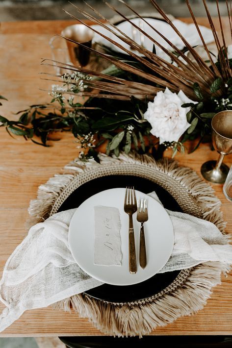 a boho western wedding place setting with a woven placemat, black and white plates, neutral linens, fronds and white blooms
