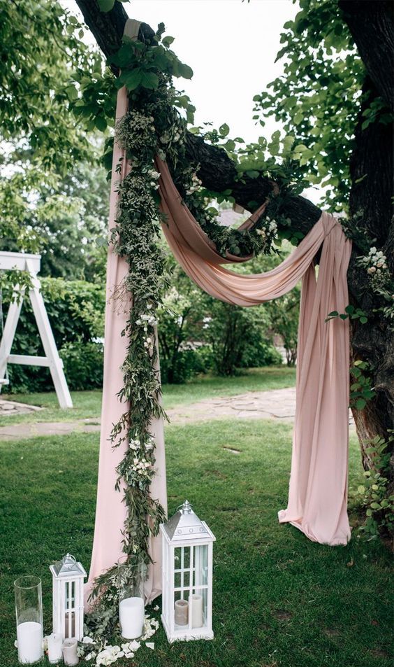 a creative wedding arch on a living tree, blush drapes, greenery and white blooms, white candle lanterns and candles
