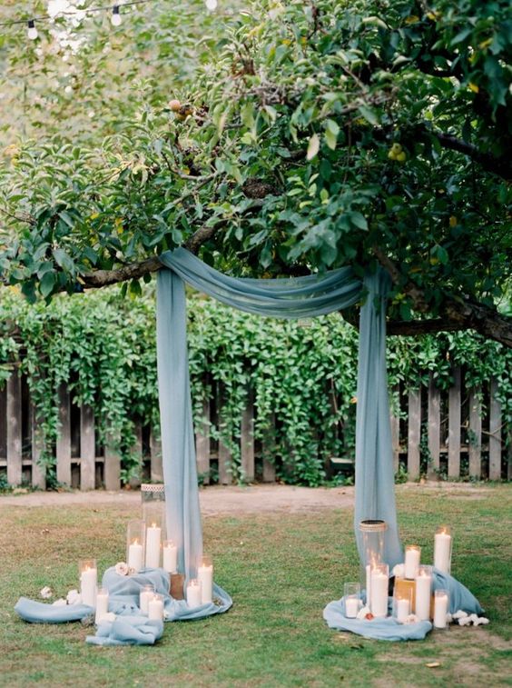 a creative wedding arch on a living tree, with dusty blue drapes, pillar candles is a very cool and chic idea