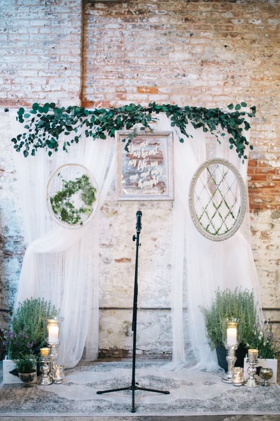 a creative wedding backdrop with white drapes, picture frames, greenery and potted blooms, pillar candles and a quote frame is amazing