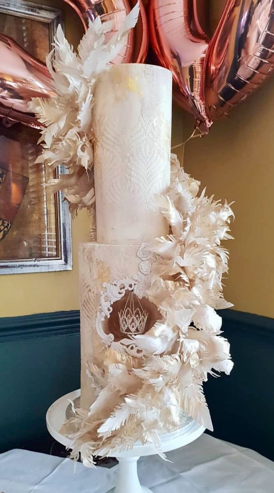 a dramatic neutral wedding cake with patterns and gold touches and a hanging crown inside plus neutral feather swirls around