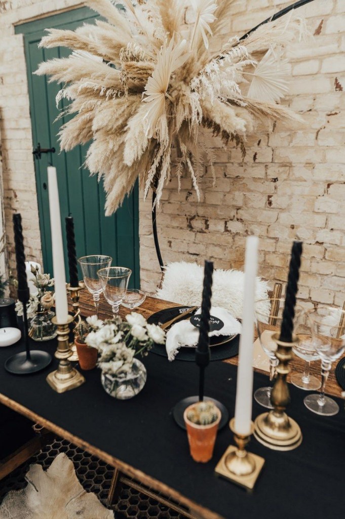 a bold western wedding tablescape with a black runner and black and white candles, black placemats and gilded touches plus pampas grass over the space