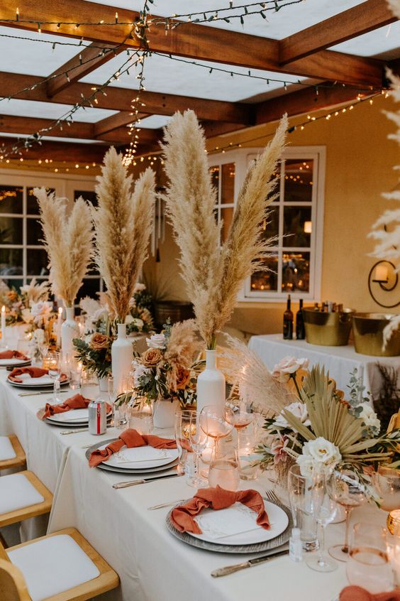 a bright and catchy western wedding tablescape with pampas grass, fronds, neutral and fall-colored blooms, lights, rust napkins