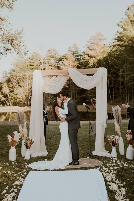 a modern boho wedding backdrop with neutral drapes, white vases with pampas grass and fronds is a lovely idea for a wedding