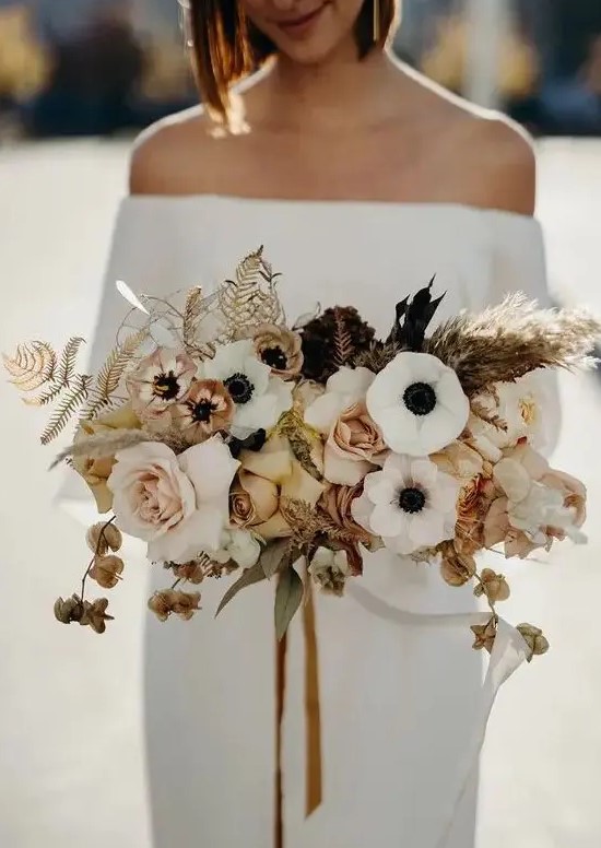 a stylish modern wedding bouquet of white anemones, blush and rust roses, dried blooms, leaves and grasses and mustard ribbons