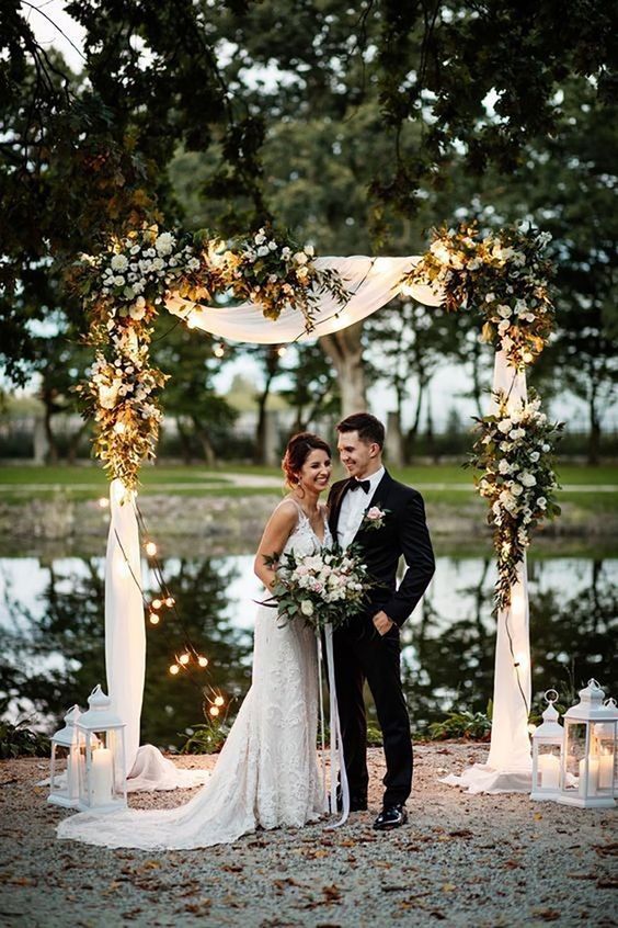 a neutral wedding arch with white drapes, white blooms and greenery, white candle lanterns is a lovely and timeless idea for a wedding