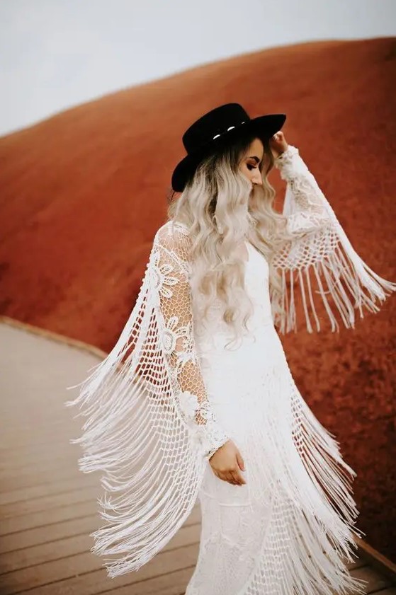 a western bridal outfit with a boho lace wedding dress with long fringe and a black hat for a bold and contrasting touch