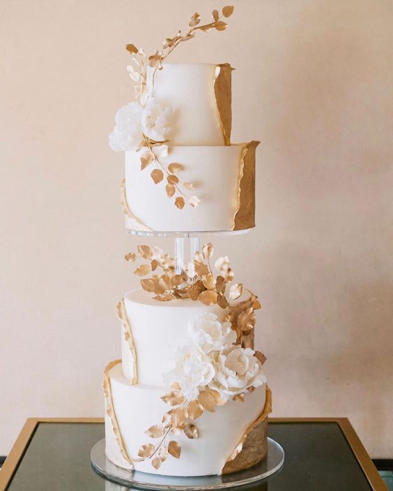 a white and gold wedding cake with a clear stand in the center, with sugar blooms and leaves and gold leaf swirls around it