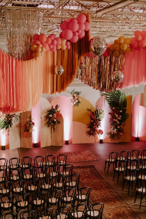 a jaw dropping colorful wedding ceremony space with bright backdrops, blooms and fronds, colorful streamers over the space and cool chairs