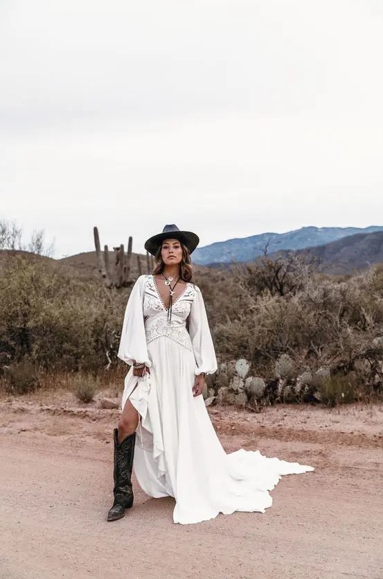 a western bridal outfit with a gorgeous boho lace wedding dress with a train, a black hat, tall black cowgirl boots and statement accessories
