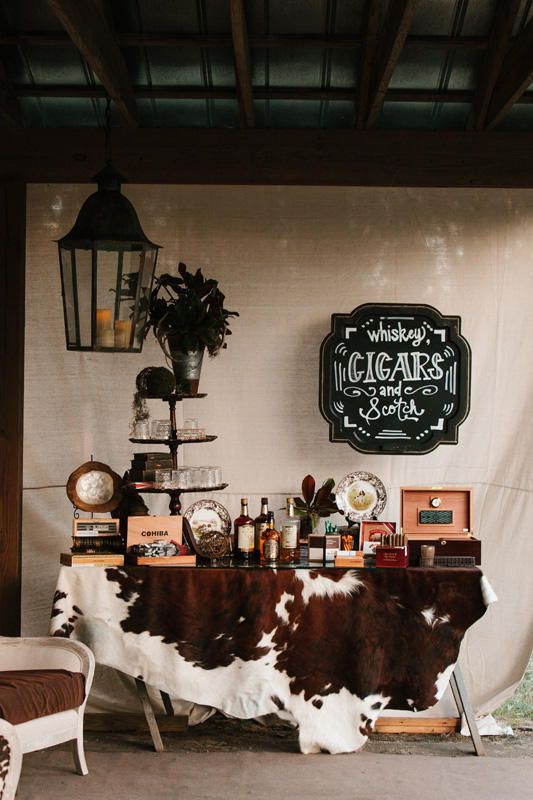 a western wedding bar with a cowhide rug, some boxes with cigars, bottles of alcohol, a sign and potted greenery is col