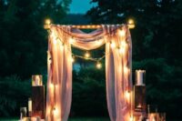 27 a pretty romantic wedding arch with pink drapes, lights, pillar candles, white petals on the ground for a night wedding ceremony