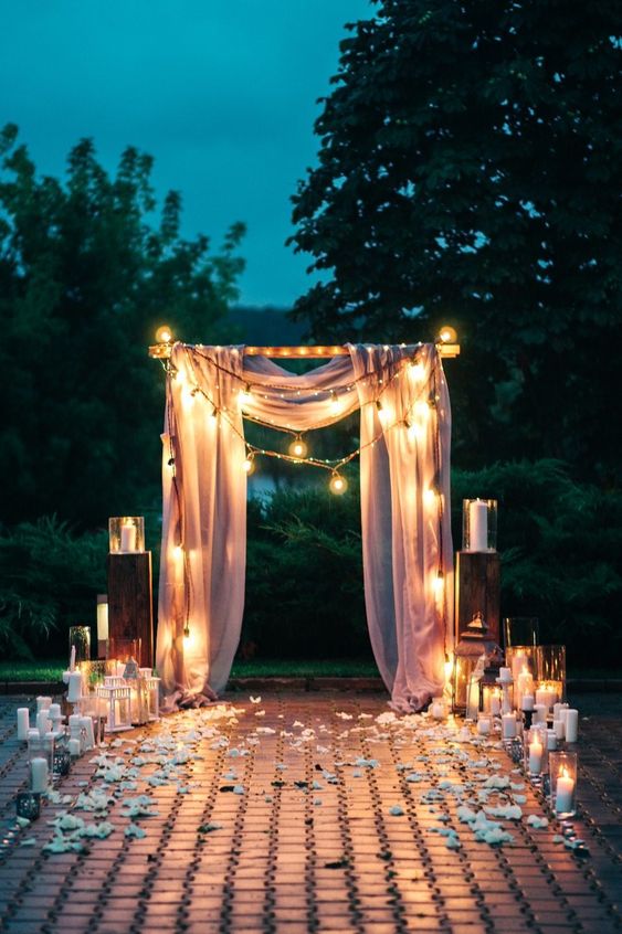 a pretty romantic wedding arch with pink drapes, lights, pillar candles, white petals on the ground for a night wedding ceremony