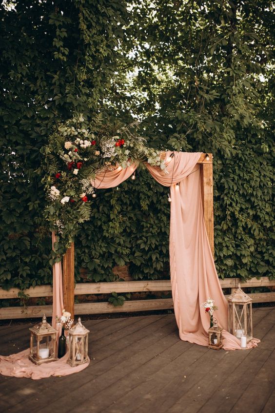 a refined wedding arch with pink fabric, greenery, red and white blooms and bulbs, candle lanterns and pillar candles