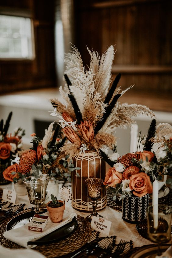 a western wedding centerpiece of patterned vases, rust and orange blooms, dried grasses and candles is amazing for a boho wedding