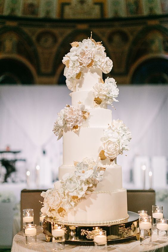 a white wedding cake with white sugar blooms and gold leaf swirls around it is a very chic and beautiful idea for a formal wedding