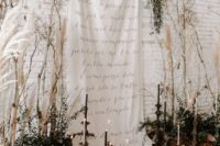 30 a romantic wedding backdrop of a white drape with quotes, greenery and dried branches, tall and thin candles and fall leaves