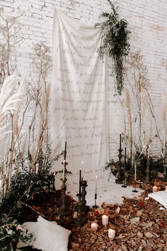 a romantic wedding backdrop of a white drape with quotes, greenery and dried branches, tall and thin candles and fall leaves