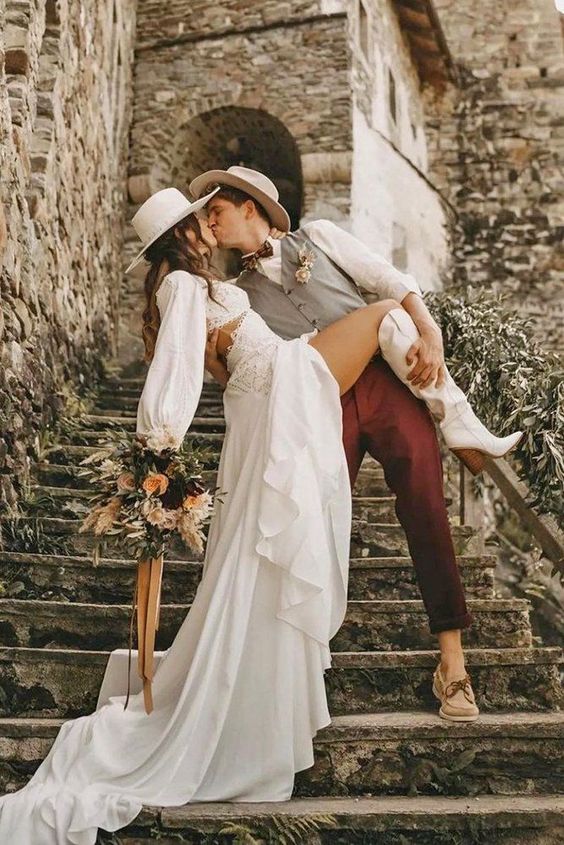 a chic western bridal look with a boho lace crop top with long sleeves, a high waisted skirt with a train, white cowboy boots and a neutral hat