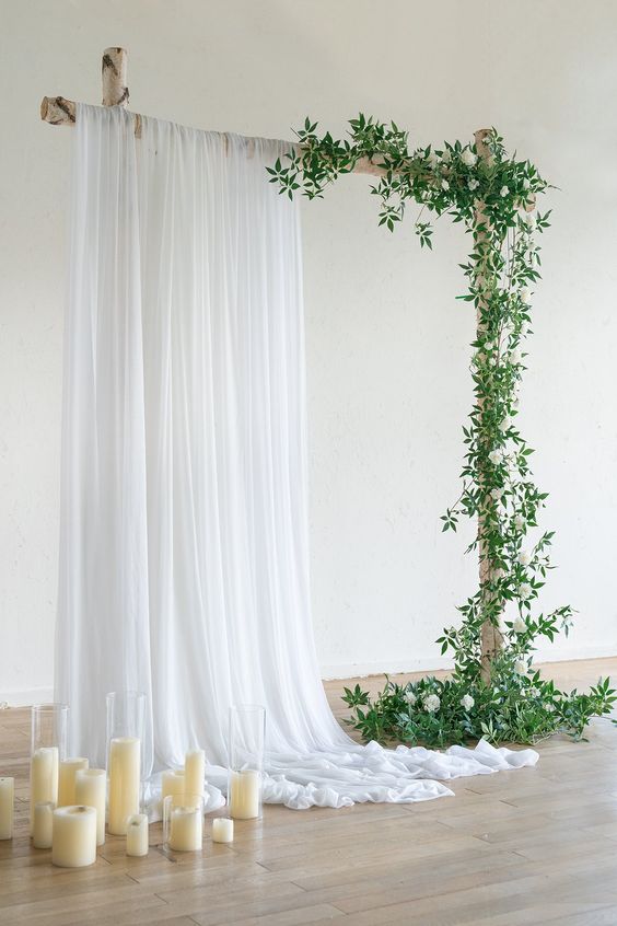 a simple and classy wedding backdrop with white drapes and greenery and white blooms, pillar candles on the floor is a lovely idea