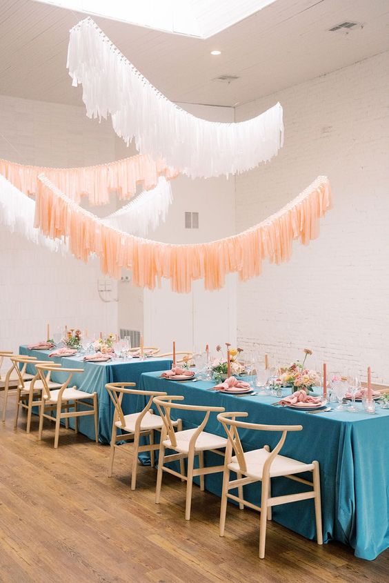a mid-century modern wedding reception space with blue tablecloths and pink blooms and candles, white and nude streamers over the space