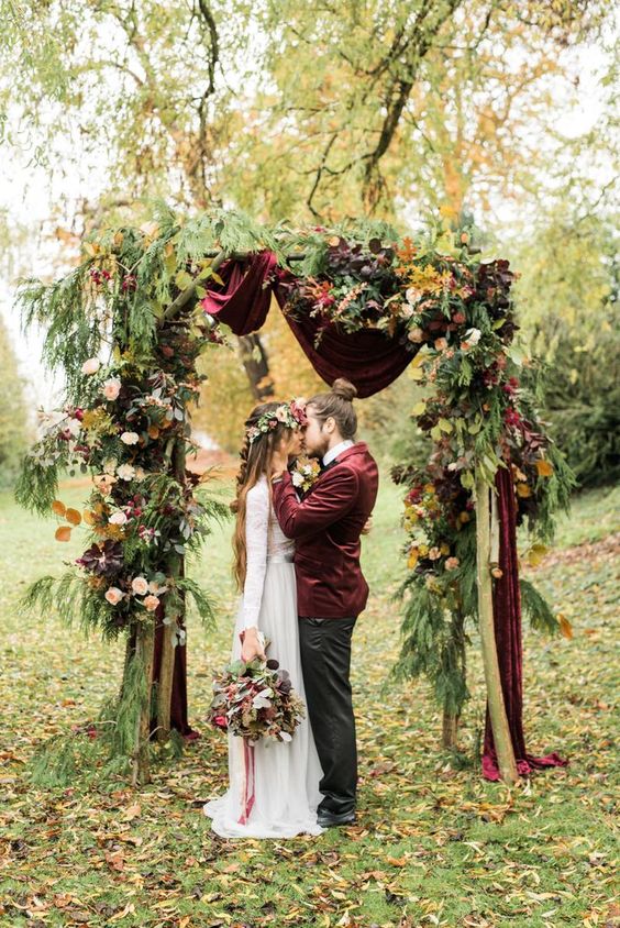 a sophisticated fall wedding arch with burgundy velvet drapes, bold blooms, fir branches and bold fall leaves for a boho wedding