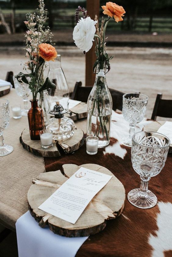 a western wedding tablescape with a burlap tablecloth, a cowhide piece, wood slice placemats and manues, bold and white blooms