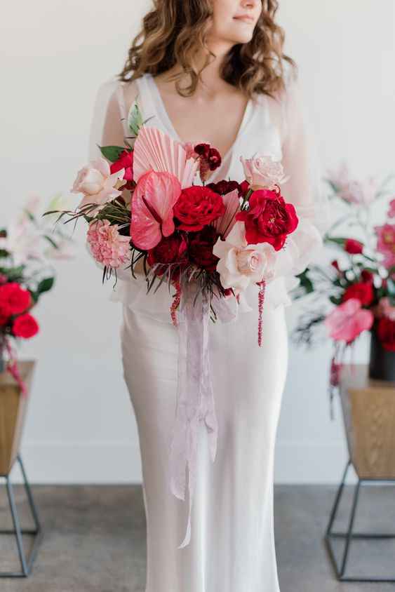 a colorful modern wedding bouquet of blush roses, red peony roses, pink fronds and anturium plus long ribbon