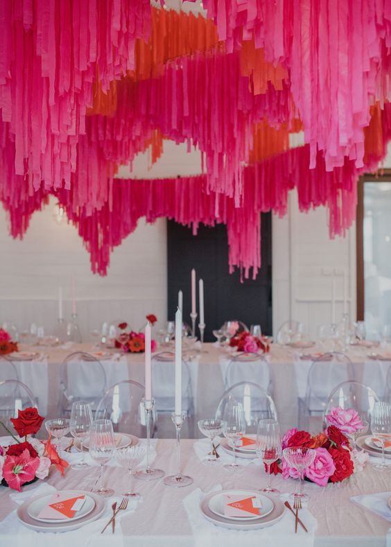 a neutral wedding reception space with white tables and pink and red blooms on the tables, hot pink and red streamers over the tables