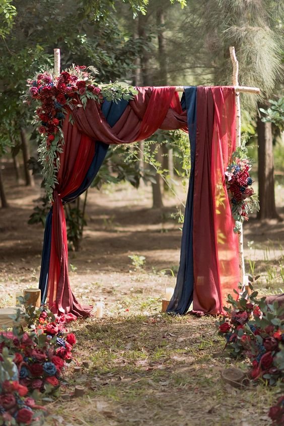 a super lush fall wedding arch with red, burgundy and navy drapes, burgundy blooms and greenery and matching blooms along the aisle