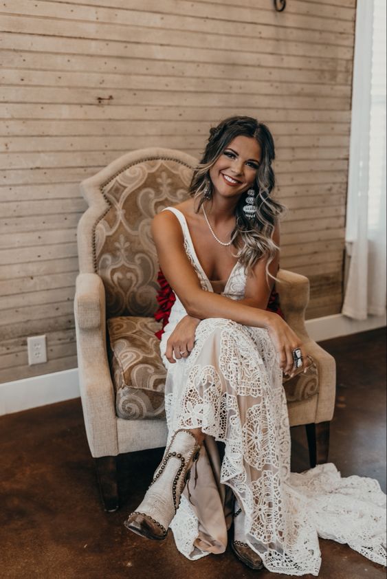 an A line boho lace wedding dress with thick straps, a covered plunging neckline, a train, neutral embellished cowbo boots and statement jewelry