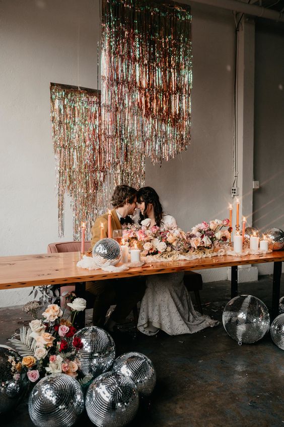 a super fun disco inspired wedding reception space with silver disco balls, blooms and silver tinsel fringe over the table