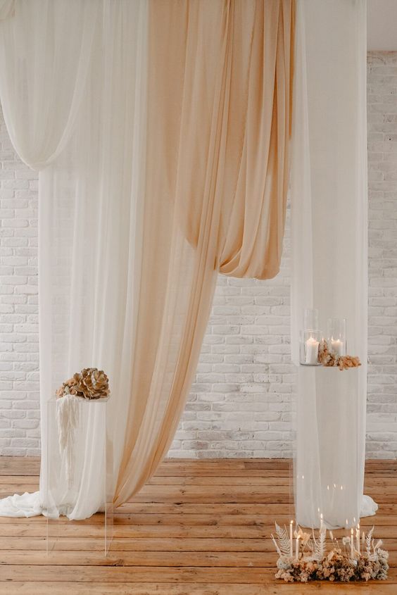 a modern wedding backdrop with neutral and peachy drapes, pillar candles, dried and fresh blooms is a beautiful and chic wedding decor idea