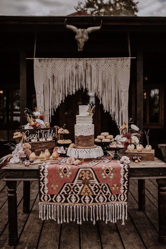 a western wedding dessert table with a woven rug, a western-style cake, cookies, pears and other sweets plus a macrame backdrop