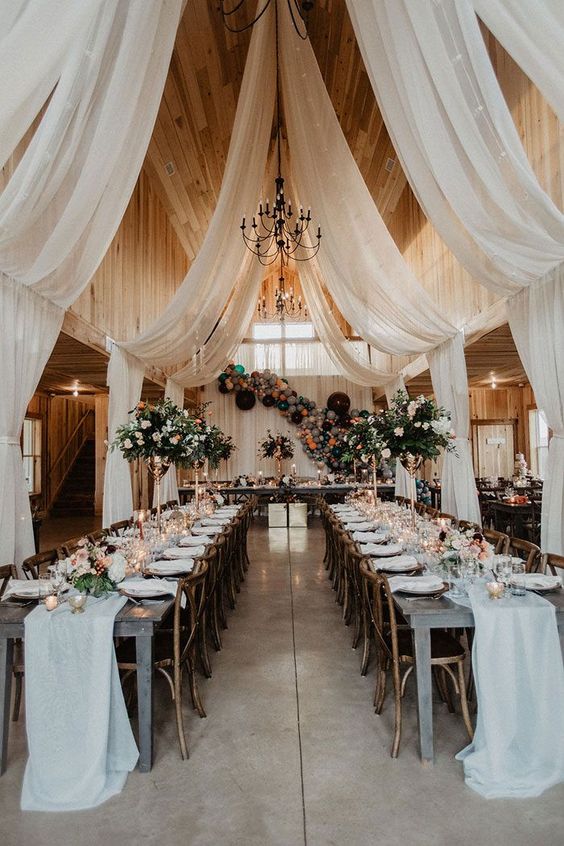 a lovely wedding reception space with white drapes and table runners, neutral blooms and greenery and candles is wow