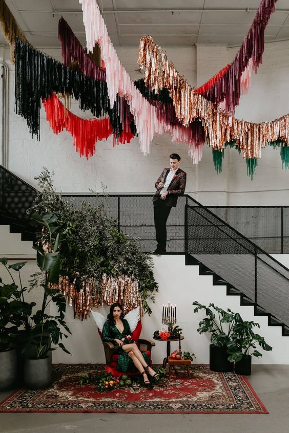 a wedding lounge with greenery, pink, red, gold and dark green streamers over the space and a small sitting space