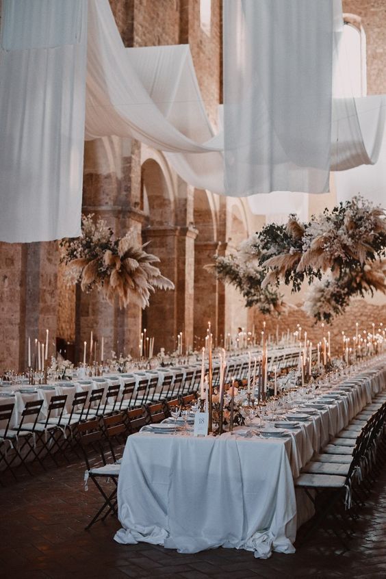 a wedding reception space with white drapes, neutral blooms, greenery and pampas grass, tall and thin candles