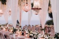 51 a white outdoor wedding reception space with an overhead greenery and white flower installation, chandeliers and white drapes and a white flower table runner