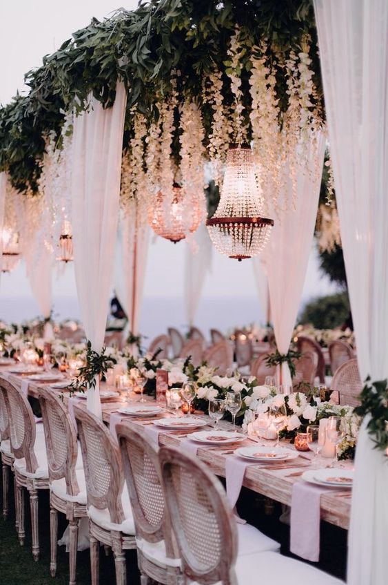 a white outdoor wedding reception space with an overhead greenery and white flower installation, chandeliers and white drapes and a white flower table runner
