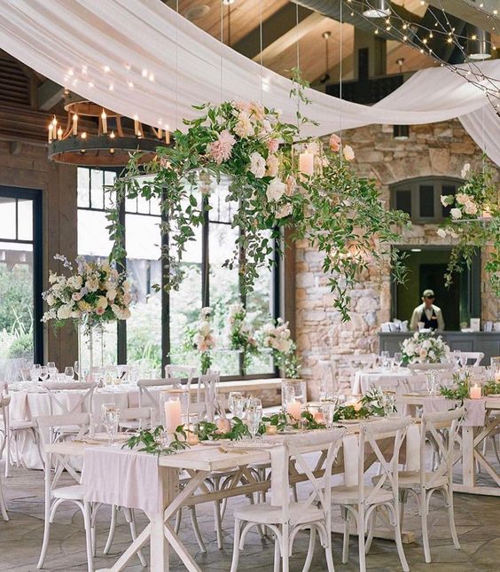 an airy and chic wedding reception space with neutral drapes and large chandeliers, white furniture, greenery and white and pink blooms