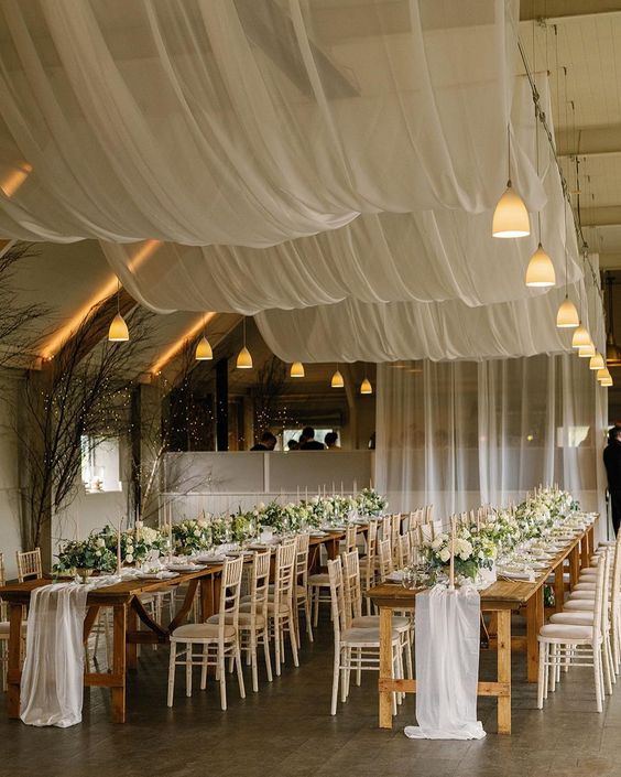 an airy wedding reception space with semi-sheer drapes over the space, white table runners, white blooms and greenery