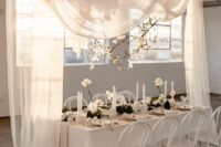 54 a chic modern wedding reception space in white, with white drapes and blooming branches over the space, a white table and white roses