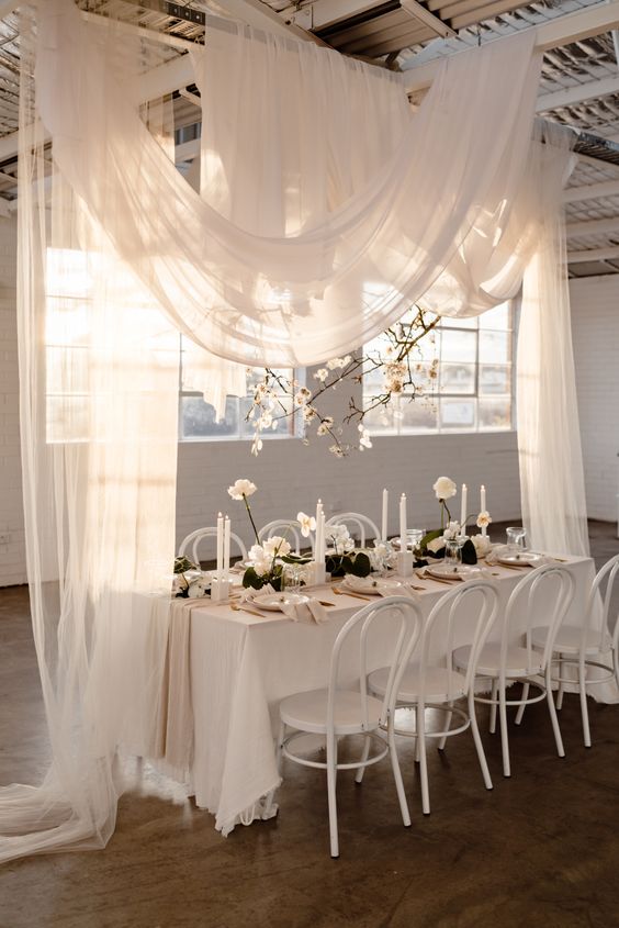 a chic modern wedding reception space in white, with white drapes and blooming branches over the space, a white table and white roses