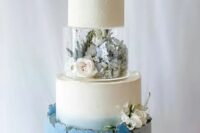 a blue and white wedding cake with a clear tier filled with fresh neutral blooms, with a texture and a rough edge plus blooms on top