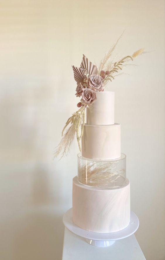 a blush wedding cake with a pattern, a clear acrylic separator with some dried grasses, dusty pink fronds and blooms plus grass on top