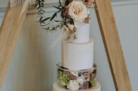 a boho neutral wedding cake with a clear acrylic separator filled with blooms and greenery, some blooms, dried grasses and a frond on top
