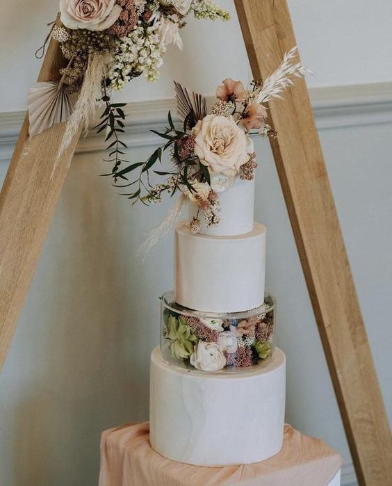 a boho neutral wedding cake with a clear acrylic separator filled with blooms and greenery, some blooms, dried grasses and a frond on top