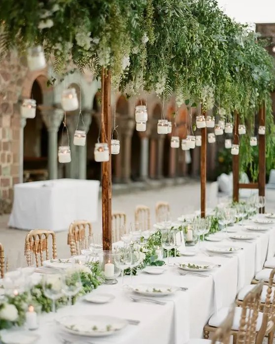 a botanical wedding venue with hanging candles, greenery, a greenery and white bloom runner and white linens is fresh and chic